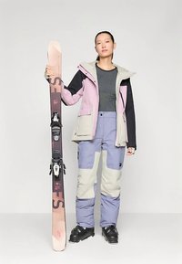 Slēpošanas apģērbs ar daudzkrāsainu jaku, kurā ir violeta, melna un pelēka paneļi, kopā ar gaisvi violetām slēpošanas biksēm, turot rozā un pelēkas slēpes.