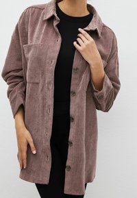 Chaqueta de camisa de pana en color malva con una tela texturizada, cierre de botones en la parte delantera, cuello con punta y bolsillo en el pecho. Mangas largas y ajuste relajado.