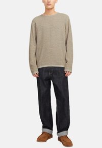 Jongere draagt een losse beige gebreide trui, donkerdenim jeans met opgerolde pijpen en bruine suède veterschoenen, staande tegen een effen achtergrond.