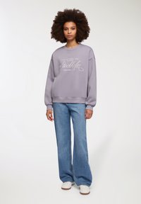 Fioletowa bluza z napisem "Stockholm Leisure Club" w białym kolorze, połączona z jasnoniebieskimi dzwonami i białymi trampkami. Gładka faktura, luźny krój.