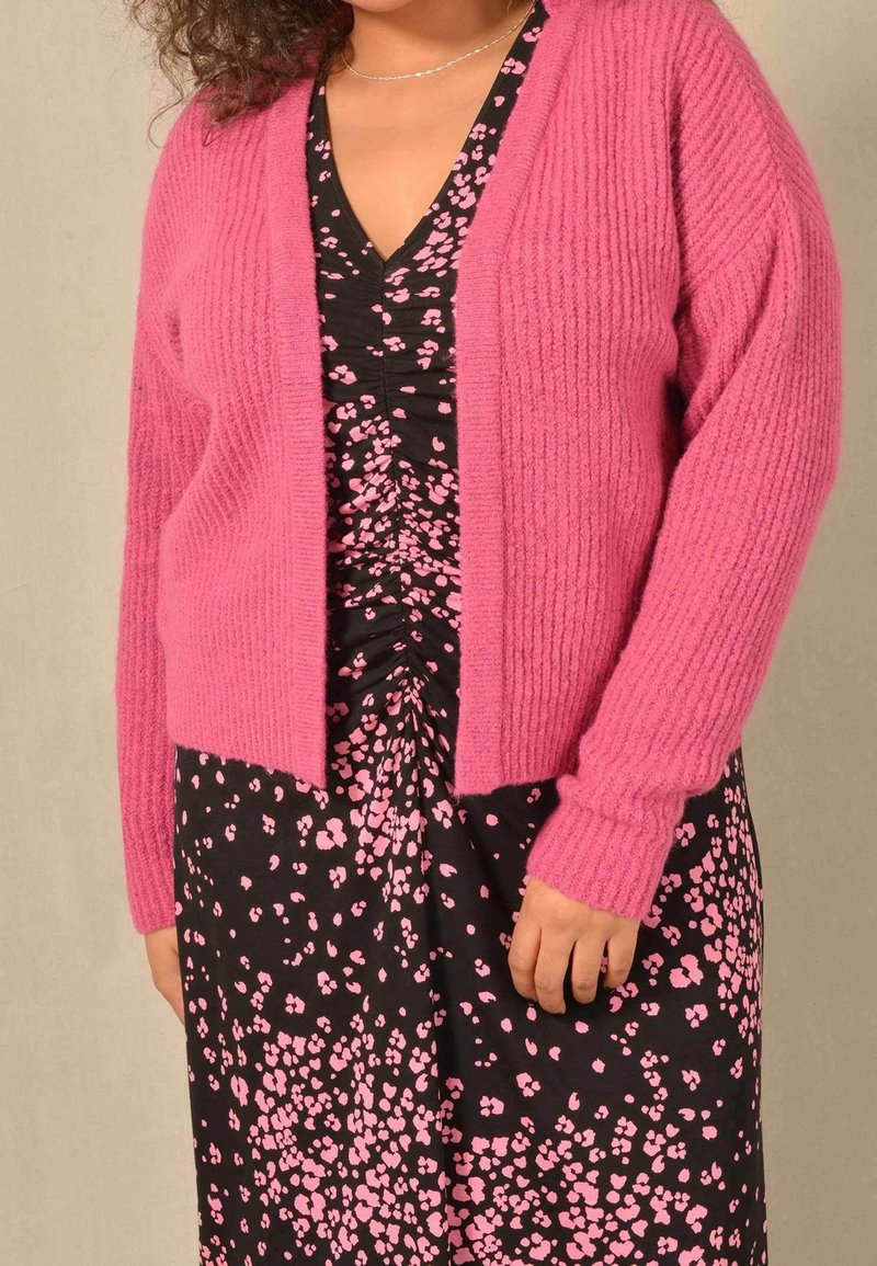 Růžový žebrovaný kardigan s pohodlným střihem a otevřeným předním dílem, doplněný černými šaty s růžovým květinovým vzorem. Měkký, texturovaný materiál.