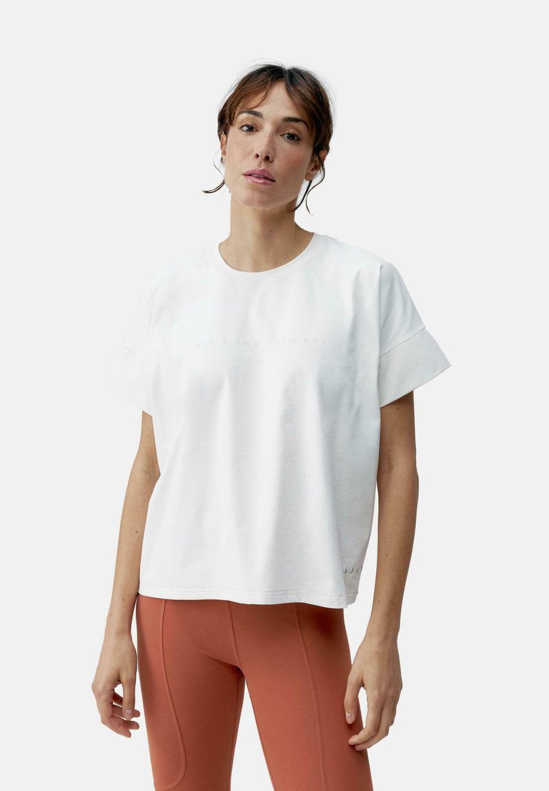 Balta trumpa rankovėmis marškinėliai su apvalia iškirpte, puoštos subtiliu tekstiniu dizainu. Medžiaga atrodo švelni, derinama su oranžinėmis leginsais.