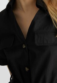 Camicia nera con bottoni, maniche corte, presenta un colletto a scollo a V, due tasche sul petto e bottoni marroni. Tessuto liscio e leggero.
