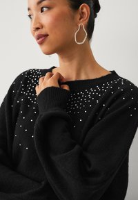 Sort sweater med lange ærmer og en afslappet pasform, pyntet med spredte sølvfarvede rhinsten på skuldrene. Struktureret strikstof.