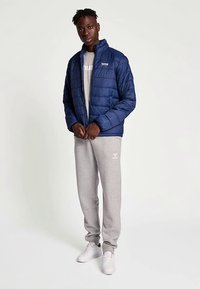 Veste matelassée bleu marine avec motif losange, fermeture éclair et col montant. Portée par-dessus un sweat-shirt gris clair et un pantalon de survêtement gris.
