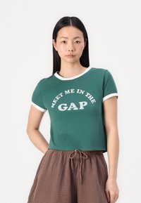 Green short-sleeve T-shirt with white trim, featuring "MEET ME IN THE GAP" graphic in a playful font. Soft cotton material, cropped fit.