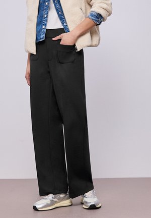 Personne portant un pantalon noir à jambes larges avec poches avant, baskets argentées, chemise en denim, veste en polaire crème, main dans la poche, debout sur un sol clair.