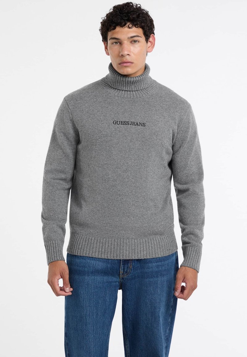 Maglione grigio con un alto collo a costine, polsini e orlo a costine. Presenta il logo nero "GUESS JEANS" sul petto. Abbinato a jeans blu.