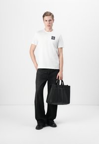 Vit t-shirt med en svart ficklogotyp, kombinerad med mörka vida jeans och en strukturerad handväska i svart läder. Svarta slip-on skor.