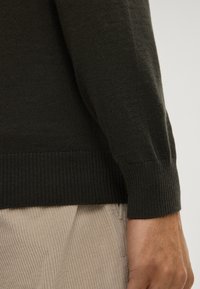 Dunkelgrüner Strickpullover im Nahaufnahme, mit gerippten Bündchen und Saum, kombiniert mit hellbeigen Cordhosen. Strukturierter und figurbetonter Schnitt.