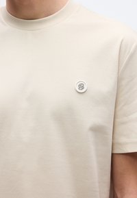 Μπεζ βαμβακερό t-shirt με λαιμόκοψη στρογγυλή, που διαθέτει μικρή λευκή κουμπί στη �αριστερή πλευρά του στήθους. Λεία υφή, μινιμαλιστικός σχεδιασμός.