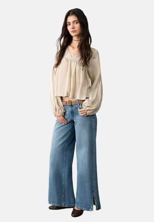 Junge Frau mit langen dunklen Haaren, die eine lockere beige Bluse, weite blaue Jeans mit Seitenschlitzen, einen braunen Gürtel und dunkle Schuhe trägt, stehend.