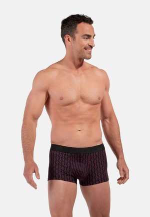 Lächelnder, oberkörperfreier Mann, der schwarze Boxer-Shorts mit einem roten geometrischen Muster trägt und vor einem schlichten hellen Hintergrund steht.