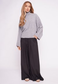 Szary dzianinowy sweter z golfem, z prążkowanymi mankietami i dołem, w połączeniu z czarnymi plisowanymi spodniami o szerokich nogawkach oraz beżowymi butami na podwyższeniu.