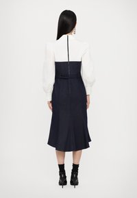 Denim jurk met een witte top, heeft een hoge kraag, lange pofmouwen en een uitlopende rok, afgewerkt met een rits aan de achterkant en een riem.