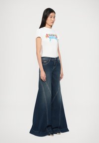 Biele tričko s grafickou potlačou, skombinované s tmavými džínsami so širokými nohavicami, ktoré majú kontrastné švy a zavonok rozšírený lem.