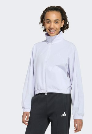Giovane donna sorridente che indossa una giacca bianca con zip e pantaloni neri Adidas con un logo bianco sulla gamba sinistra.