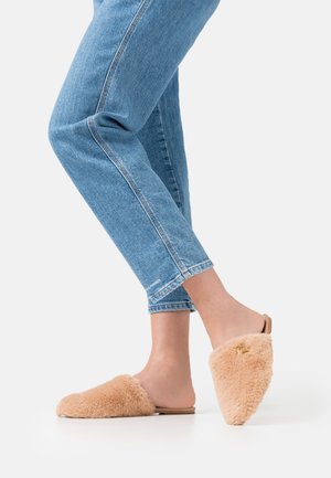 Pantoufles beige floues à enfiler avec un design pointu, associées à un jean en denim bleu retroussé. Les pantoufles présentent une texture douce et des petits éléments métalliques discrets.