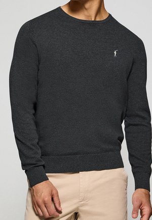 Mann trägt dunkelgrauen Pullover mit kleinem weißem Golf-Logo auf der Brust, kombiniert mit beiger Hose.