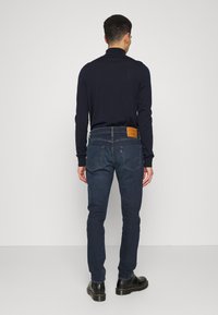 Camisa azul-marinha de mangas longas combinada com jeans azul-escuro. Os jeans possuem um patch de couro na cintura e detalhes de costura subtis. Sapatos pretos completam o look.