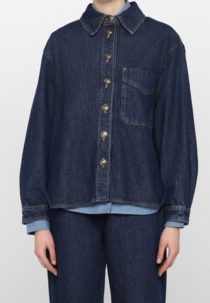 Osoba ubrana w ciemnoniebieską kurtkę jeansową na guziki z przednią kieszenią na piersi oraz dopasowane spodnie jeansowe na jednolitym tle.