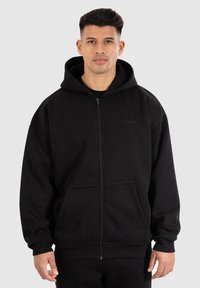 Uomo che indossa un hoodie nero con cerniera, dotato di tasche e cappuccio, in piedi su uno sfondo grigio chiaro uniforme.