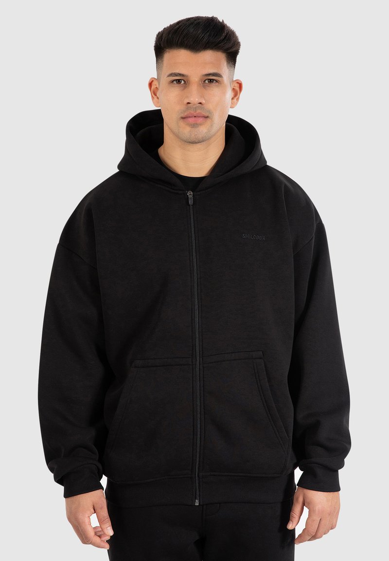 Uomo che indossa un hoodie nero con cerniera, dotato di tasche e cappuccio, in piedi su uno sfondo grigio chiaro uniforme.