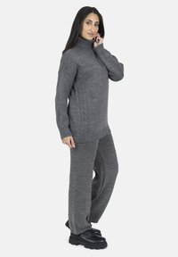 Maglione grigio lavorato a maglia con un collo alto, caratterizzato da un design a treccia e dettagli a coste. Abbinato a pantaloni grigi a gamba larga e stivali neri chunky.
