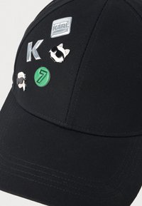 Gorra de béisbol negra con tela texturizada, que presenta múltiples pines decorativos: logo plateado "KARL", número verde "7" y un gato de dibujos animados con gafas de sol.