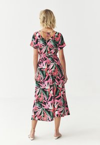 Robe midi florale pr�ésentant un motif de feuilles roses et vertes vibrantes, des manches courtes, un détail à nouer à l'arrière et un ourlet légèrement évasé.