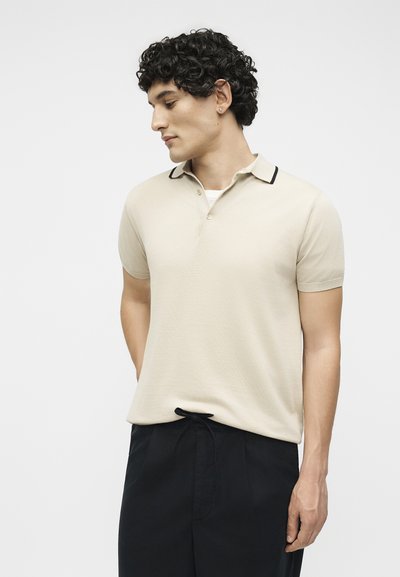 Νεαρός άντρας με σγουρά μαλλιά, που φοράει ένα μπεζ Polo με κοντά μανίκια και μαύρο παντελόνι, κοιτάζει προς τα αριστερά σε ένα απλό φόντο.