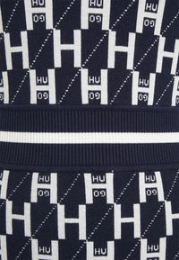 Pull en tricot présentant un motif géométrique bleu marine et blanc avec des motifs répétés de "H" et "HU" ; taille en côtes bleu marine et texture.