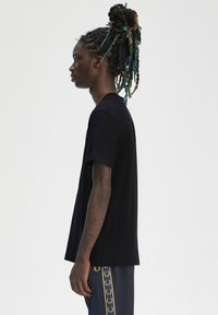 T-shirt nero a maniche corte, realizzato in cotone morbido. Presenta un collo rotondo e una vestibilità rilassata. Indossato con pantaloni blu navy, decorati con un fregio logo dorato.