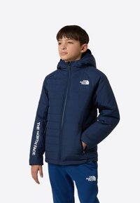 Laivastonsininen eristetty takki hupulla, jossa on vetoketju edessä ja valkoiset logoyksityiskohdat rinnassa ja hihassa. Sileäpintainen kangas.