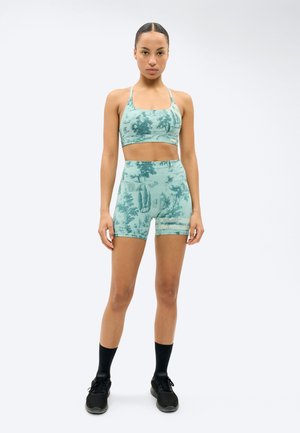 Nainen seisoo vedenvihreässä urheilurintaliivissä ja yhteen sopivissa korkeavyötäröisissä pyöräilyshortseissa, mustat keskipitkät sukat ja mustat lenkkarit jalassa.