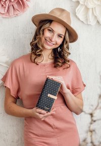 Black wallet with white polka dots and tan accents, featuring a snap closure. The background includes textured paper flowers in soft colors.