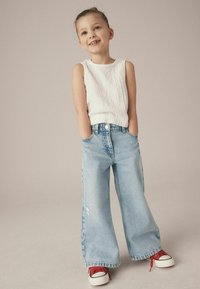 Vit texturerad ärmlös topp, ljusblå vida jeans med slitna detaljer och röda sneakers med svarta accenter.