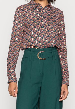 Woman wearing long-sleeve button-up blouse with orange and cream fan pattern tucked into high-waisted dark green pants with a wide belt.