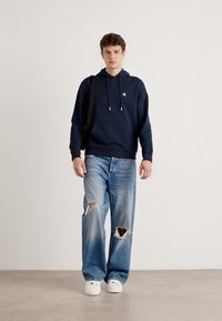 Felpa navy con cordini, abbinata a jeans strappati di un azzurro chiaro. Sneakers bianche completano l'outfit informale. Il tessuto sembra morbido e rilassato.