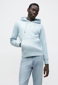 Jasnoniebieska bluza z kapturem, kieszenią z przodu oraz logo. W połączeniu z jasnoniebieskimi dżinsami. Gładka faktura, casualowy design.