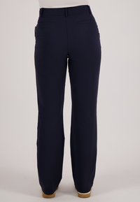 Persoon die donkerblauwe, recht gesneden broek en beige schoenen draagt, staand tegen een effen lichte achtergrond.