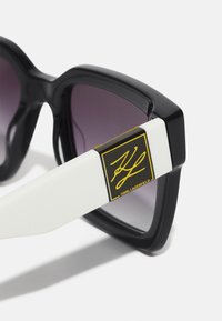Lunettes de soleil noires surdimensionnées avec des verres dégradés violets, monture blanche présentant une plaque logo dorée "Karl Lagerfeld".