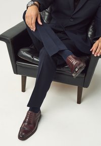 Chaussures de ville en cuir marron avec des détails brogue, portées avec un pantalon bleu marine foncé et des chaussettes assorties, assis sur une chaise en cuir noir.