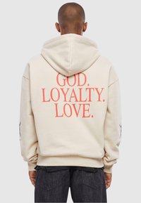 Beige hoodie med en stor, fed grafik i koralfarvet tekst, som siger "GUD. LOYALITET. KÆRLIGHED." Har også et designelement på venstre ærme.