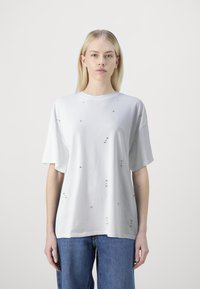 Camiseta blanca de corte oversized hecha de algodón, con adornos dispersos. Mangas cortas y diseño de cuello redondo. Sencilla y texturizada.
