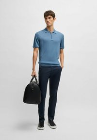 Pikétröja i ljusblått med korta ärmar och dragkedja, parat med mörkblå byxor. Modellen håller en svart läderväska och bär mörka sneakers.