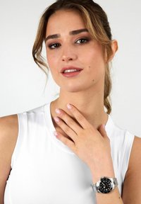 Silberne Metalluhr mit schwarzem Zifferblatt und lumineszierenden Zeigern. Getragen an einem Handgelenk mit hellrosa Nagellack vor weißem Hintergrund.