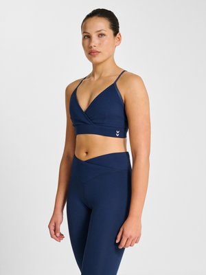 Vrouw draagt marineblauwe sportbeha en leggings met hoge taille, staand tegen een effen lichte achtergrond met een neutrale uitdrukking.