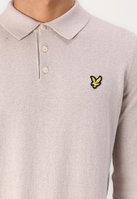 Polo shirt grigio chiaro realizzato in tessuto morbido, con colletto classico, tre bottoni e un logo aquila ricamato in giallo e nero.