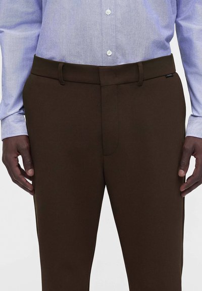 Homme portant un pantalon marron foncé avec une ceinture et une chemise bleu clair boutonnée, les mains reposant sur les côtés sur un fond uni.